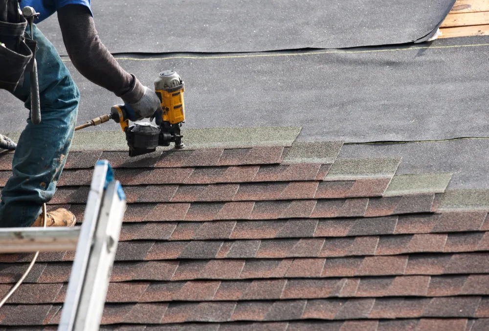 Hands installing architectural shingles with a pneumatic nailer over roof underlayment.