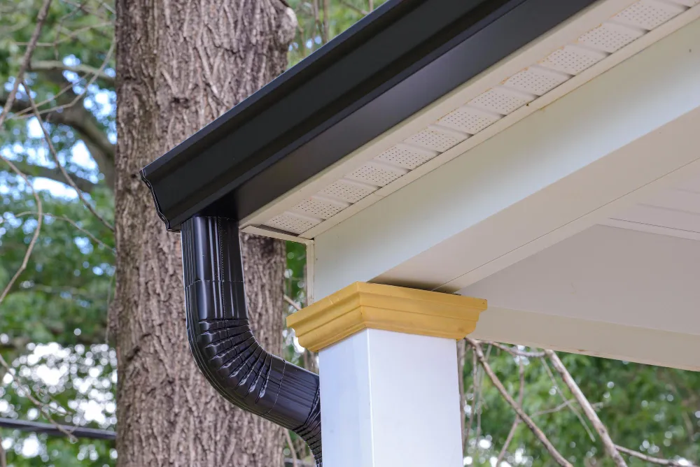 Close-up of a black seamless gutter and downspout along a home eave with vented soffit.