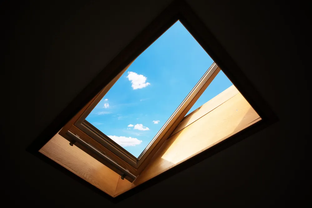 Open wood-framed skylight roof window viewed from inside, showing blue sky and clouds.