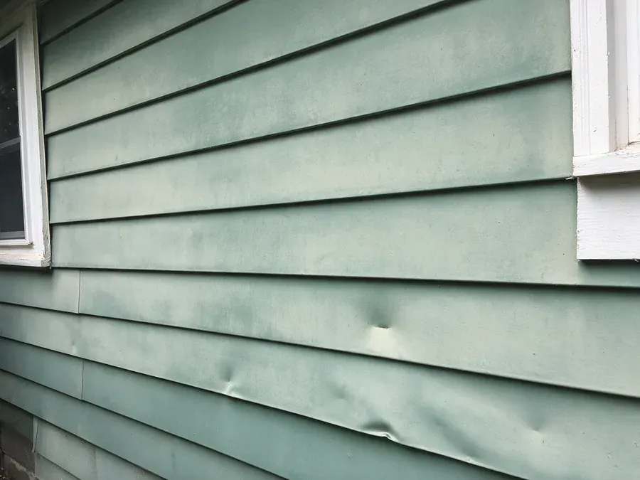 Faded green aluminum siding with dents and a bent lower edge beside a white window trim.