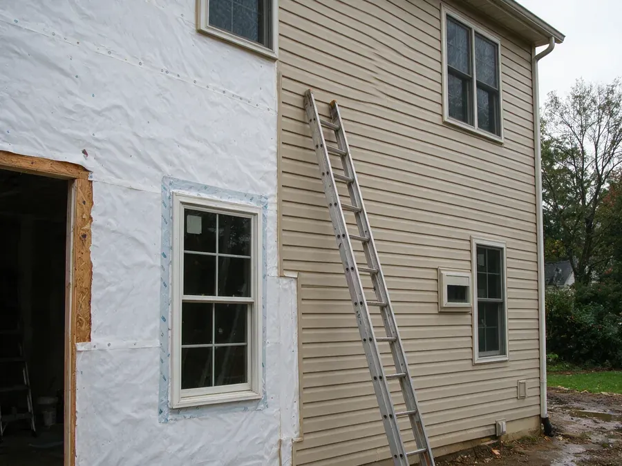 Siding replacement in progress, with house wrap and taped window flashing on one section of wall and new tan vinyl siding on the next.