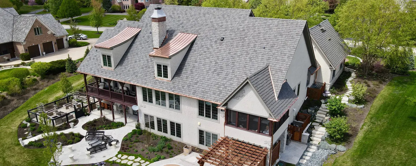 Aerial view of a stucco home with a grey shingle roof, copper-capped dormers, and landscaped gardens.