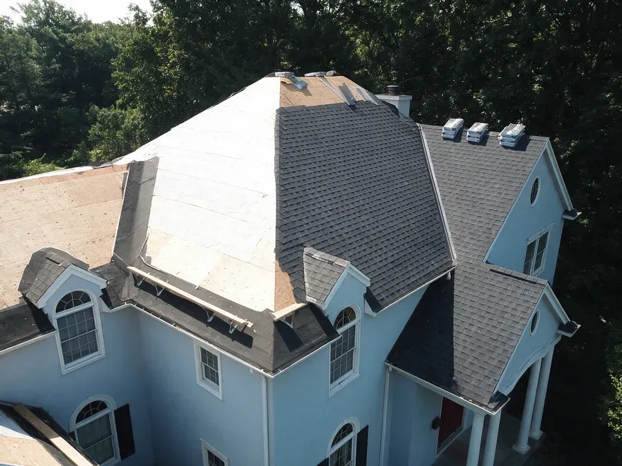 Roof replacement in progress on a large home with several roof planes, part stripped to decking and underlayment and part finished in charcoal shingles.