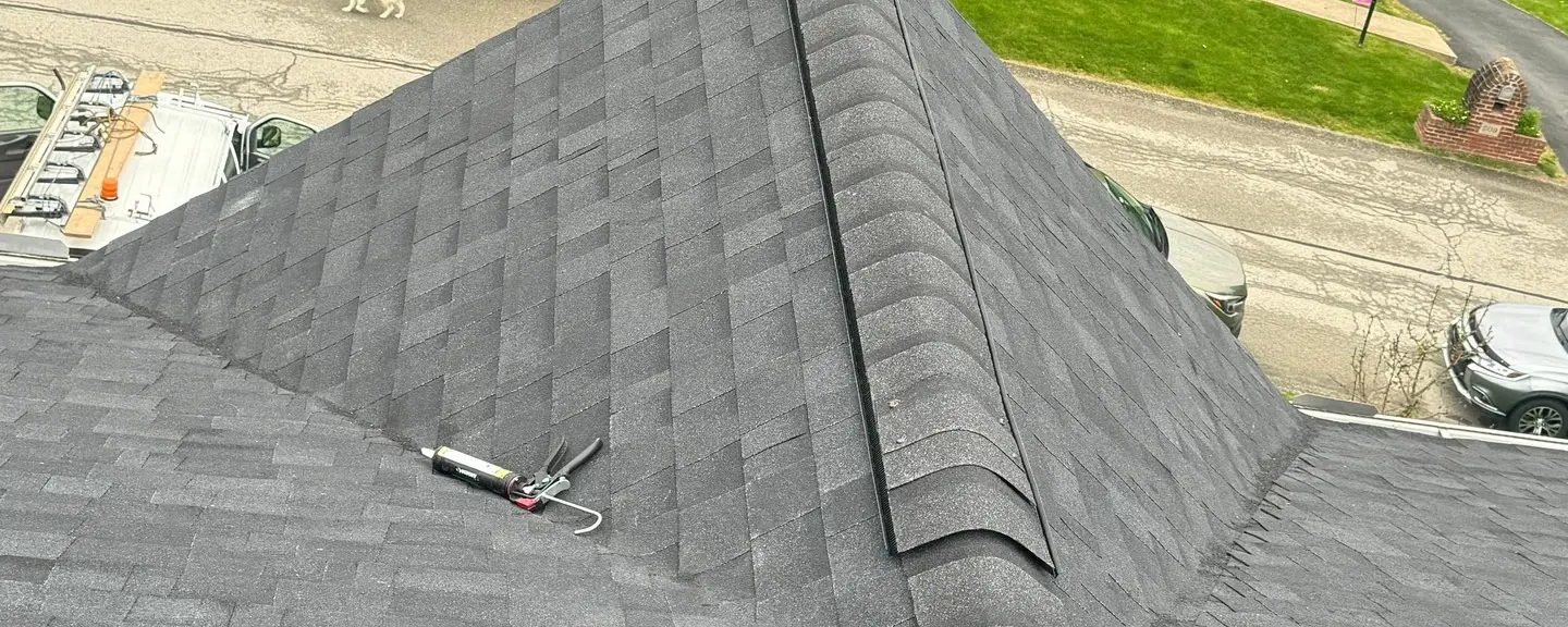 Aerial view of a finished hip roof with dark architectural asphalt shingles, roofing tools resting on the ridge, colonial home in the background.