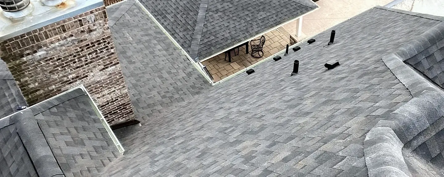 Aerial view of a complex gray architectural asphalt shingle roof with multiple hips, a brick chimney, and a covered patio nook.