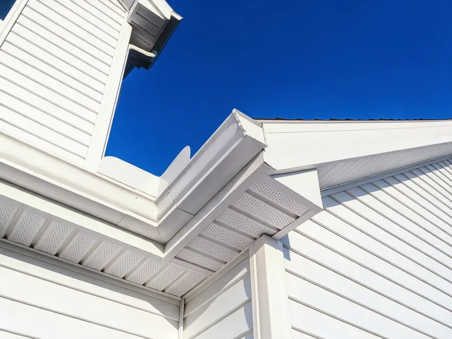 Dramatic upward view of a roofline corner with new white vinyl siding against a vivid blue sky.